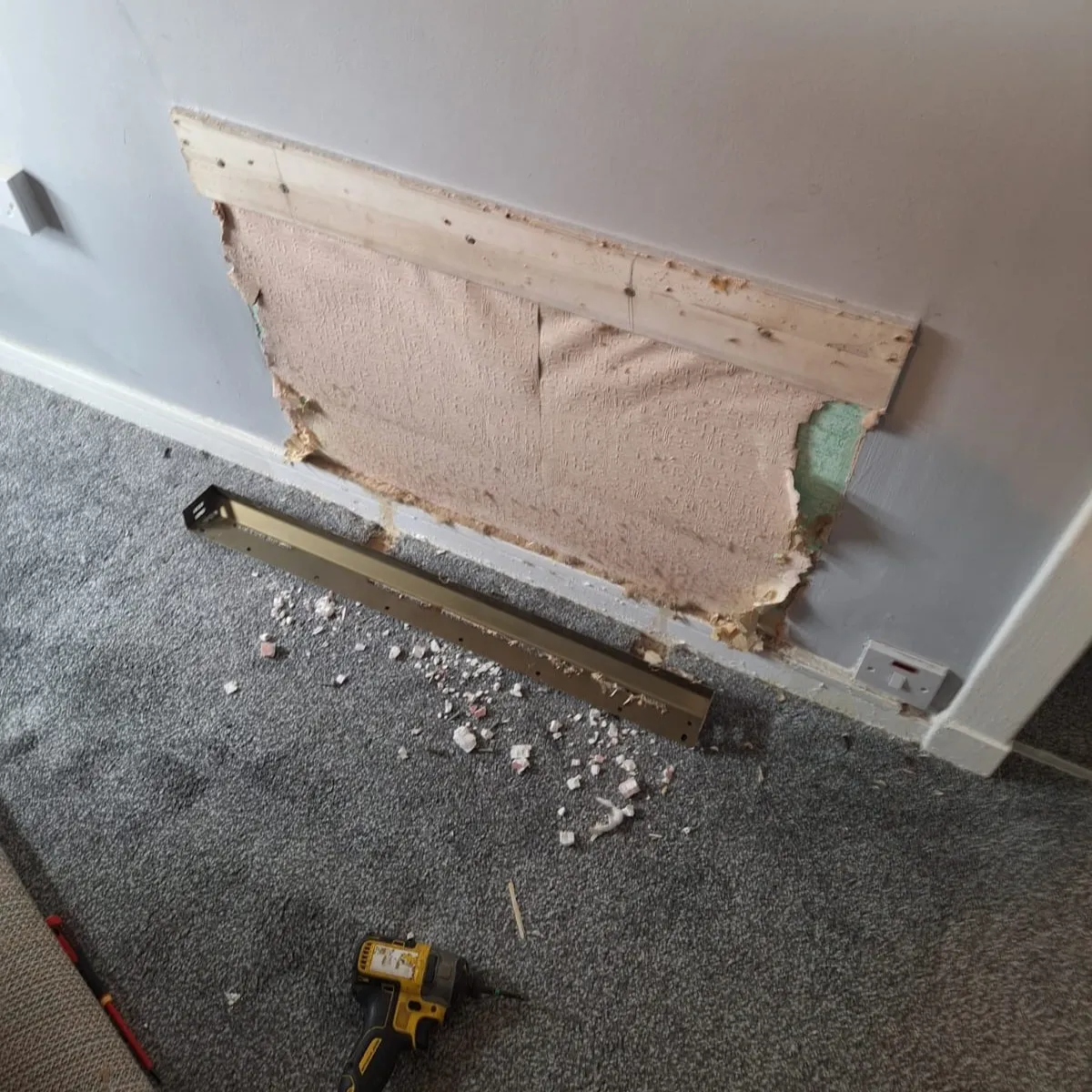 PW-19-Electri-Storage-High-Heat-Retention-Installation-a wooden access panel or patch screwed into a cutout hole in a lower drywall wall, surrounded by unpainted joint compound or plaster patch work.